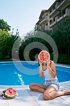 Girl looks into a hole in a piece of watermelon sitting by the pool