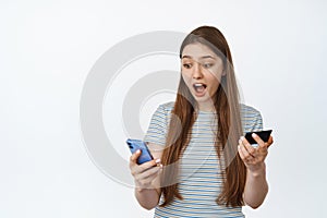 Girl looks surprised at her mobile phone, holds credit card. Concept of online shopping on smartphone, white background