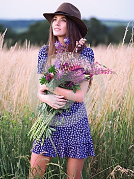 Girl with lupins in a lilac dress and a hat in a wheat field