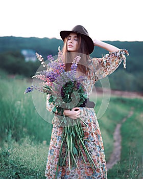 Girl with lupins in a long dress and hat at sunset