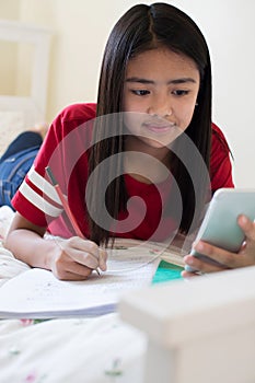 Girl Lying On Bed Using Mobile Phone To Help With Homework