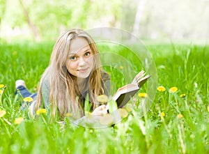 Girl lying on grass with dandelions reading a book and looking at camera