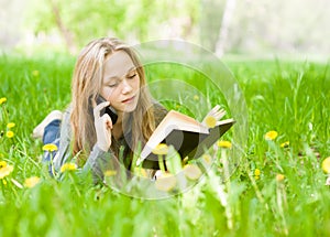 Girl lying on grass with dandelions reading a book and talking on phone