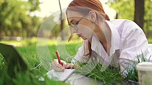 Girl lying in the grass writing something in notes and working with a laptop.