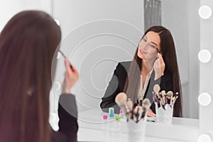 Girl, make-up artist, with long dark hair in a business suit sits at the make-up table in a bright room, office and adjusts with