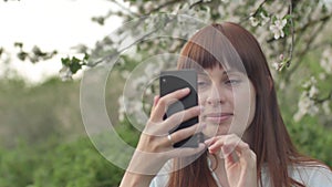 Girl makes selfie in the garden. An attractive red-haired woman smiles making selfi using a mobile phone in a cherry