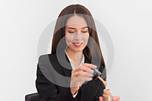 Girl, makeup artist, with long dark hair in a business suit opens a tube with concealer on a white background