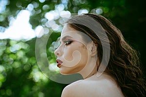 Girl with makeup face, beauty. Woman with long brunette hair on nature. Woman with curly hairstyle and bare shoulder on