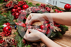 Girl making Christmas door wreath using natural fir branches, red colourful berries, wooden toys, cones and cinnamon sticks.