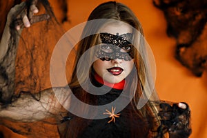A girl with a masquerade mask on her face, dressed for Halloween, looks mysteriously and holds a cobweb in her hands