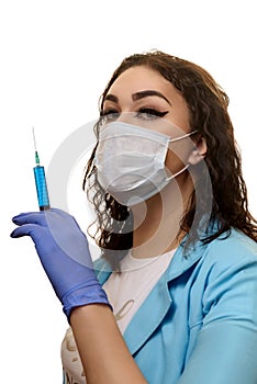 A girl in a medical mask, gloves and a robe with a syringe in his hands on a white background. Beautician, beauty specialist.