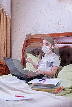 A girl in a medical mask and headphones sits on a bed with a laptop and communicates online with other conference participants. A