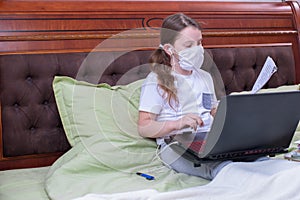 A girl in a medical mask and headphones sits on a bed with a laptop. A schoolgirl does homework, distance learning, during