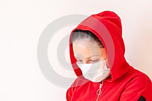 A girl in a medical mask with a hood on her head lowered her head and looks down.