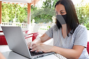 A girl in a medical mask makes a video conference through a laptop and sits outside in a cafe
