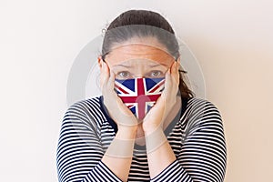 A girl in a medical mask painted like the flag of United Kingdom in fear presses her hands to her head.