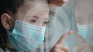 Girl in medical mask during quarantine looks sadly at her reflection in window