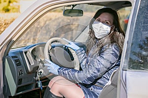 A girl in a medical mask and rubber gloves is sitting behind the wheel of a car