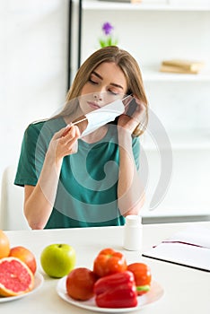 Girl in medical mask sitting at table with pills, fruits