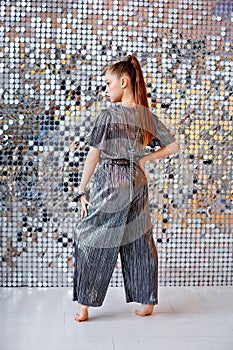 A girl in a metallic-colored jumpsuit stands with her back to the camera. It stands against the background of a shiny silver wall