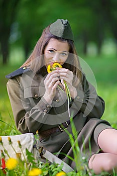 Girl in military uniform sits in a field on grass holding a bouquet