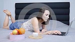 Girl millennial looking on laptop screen lying on bed in living room. Brunette young woman in shorts is chatting online on laptop