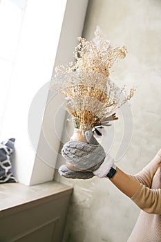 A girl in mittens holds a vase with dried flowers near the window