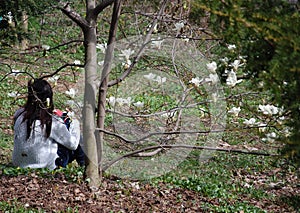 Girl with mobile phone sits under tree with blooming flowers of white magnolia