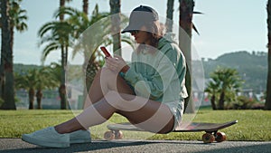 Girl with mobile sitting on skateboard in the park