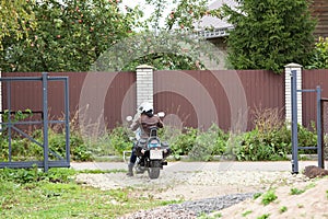 A girl on a motorcycle drives out onto the road through an open gate