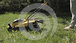 The girl is mowing an uneven lawn with yellow lawnmower barefoot