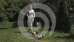 The girl is mowing an uneven lawn with yellow lawnmower barefoot