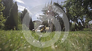 The girl is mowing an uneven lawn with yellow lawnmower barefoot