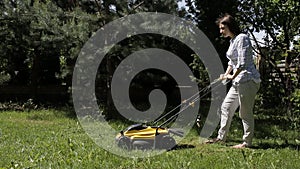 The girl is mowing an uneven lawn with yellow lawnmower barefoot