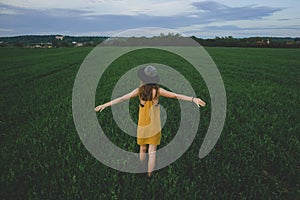 A girl in a mustard-colored dress and a hat stands in a green field