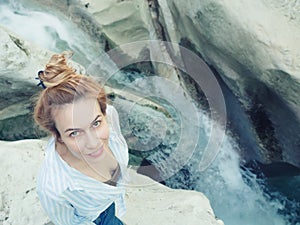 A girl with mysterious eyes looks up from below while standing on a rock near a waterfall