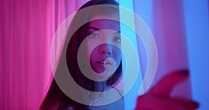 Girl in neon looks at the camera around the corner light night portrait flirts curtains pink blue