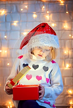 A girl in a New Year`s hat with curiosity opens a gift box from which a light shines