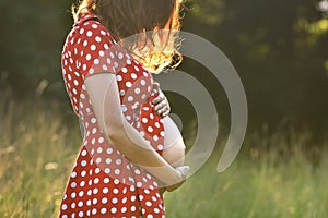 Girl in the ninth month of pregnancy in a red dress with white polka dots. Pregnant redhead woman on the meadow at sunset. 9 month