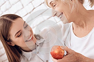 Girl is nursing elderly woman at home. They are embracing. Woman is holding apple.