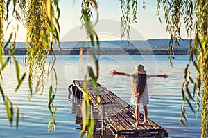 Girl on a old wooden fishing pier and willow tree enjoying beautiful sunset over the sea lake
