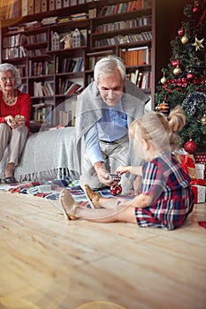 Girl is open Christmas present in front of a decorated X-mas tree with grandpa