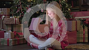 Girl opens Christmas gift sitting near Christmas tree on new year`s eve. Blonde pretty young girl opens gift magic box