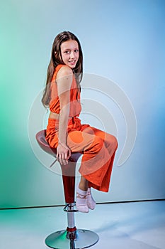 A girl in an orange suit sitting on a chair posing on a light blue and green background