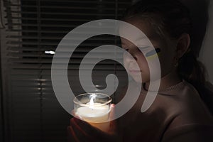 A girl with a painted flag of Ukraine on her cheek in a dark room holds a candle in her hands