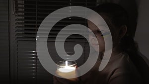 A girl with a painted flag of Ukraine on her cheek in a dark room holds a candle in her hands