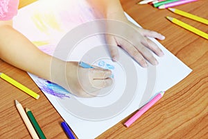 Girl painting on paper sheet with colour pencils on the wooden table at home - child kid doing drawing picture and colorful crayon