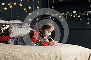 A girl in pajamas lies on the bed and writes a letter to Santa Claus or grandfather frost on the background of a Christmas tree