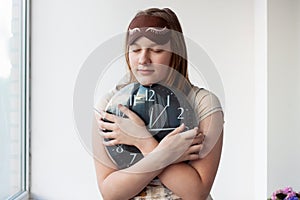 A girl in pajamas and a sleep mask with her eyes closed hugs a large black watch with white hands and numbers