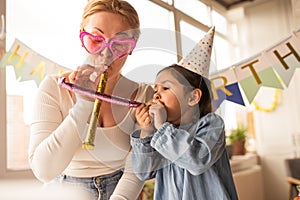 Girl in party cap blowing celebrating her birthday with her mother wearing funny glasses